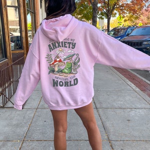 May include: Pink hoodie with a graphic of a frog and mushroom on the back. The text on the hoodie reads "Just me and my anxiety against the world."
