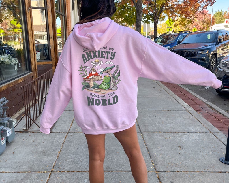 May include: Pink hoodie with a graphic of a frog and mushroom on the back. The text on the hoodie reads "Just me and my anxiety against the world."