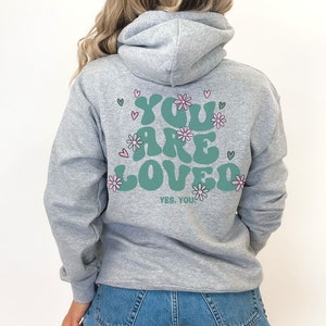 以下が含まれることがあります： 「YOU ARE LOVED」の文字がティール色で描かれたグレーのパーカー。ピンクの花とハートで装飾されています。「YES, YOU」の文字が下にあります。パーカーには、ドローストリングフードと長袖が付いています。