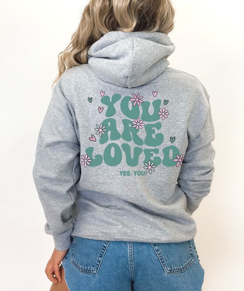以下が含まれることがあります： 「YOU ARE LOVED」の文字がティール色で描かれたグレーのパーカー。ピンクの花とハートで装飾されています。「YES, YOU」の文字が下にあります。パーカーには、ドローストリングフードと長袖が付いています。