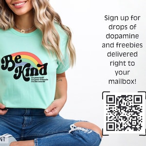 Puede incluir: Una camiseta verde menta con un dise&ntilde;o de arco&iacute;ris retro y las palabras "Be Kind" en negrita negra. La camiseta tambi&eacute;n incluye el texto "To yourself, To your friends, To the world". La camiseta se combina con unos vaqueros azules.