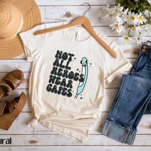 May include: A beige t-shirt with a graphic of a blue syringe with a smiling face and the text "Not all heroes wear capes".