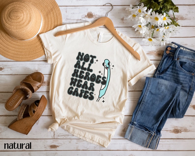 May include: A beige t-shirt with a graphic of a blue syringe with a smiling face and the text "Not all heroes wear capes".