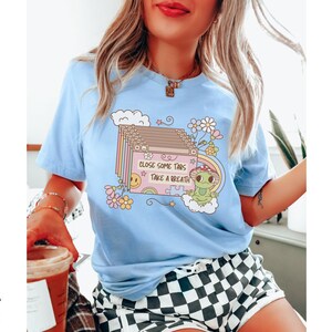May include: A light blue t-shirt with a graphic of a cartoon frog and the text "Close some tabs, take a breath". The graphic is surrounded by flowers and clouds.