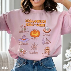 May include: A light pink sweatshirt with the text "HALLOWEEN SELF-CARE" in black lettering. The sweatshirt features a variety of Halloween-themed illustrations, including a pumpkin, a witch's hat, a spiderweb, and a tombstone.