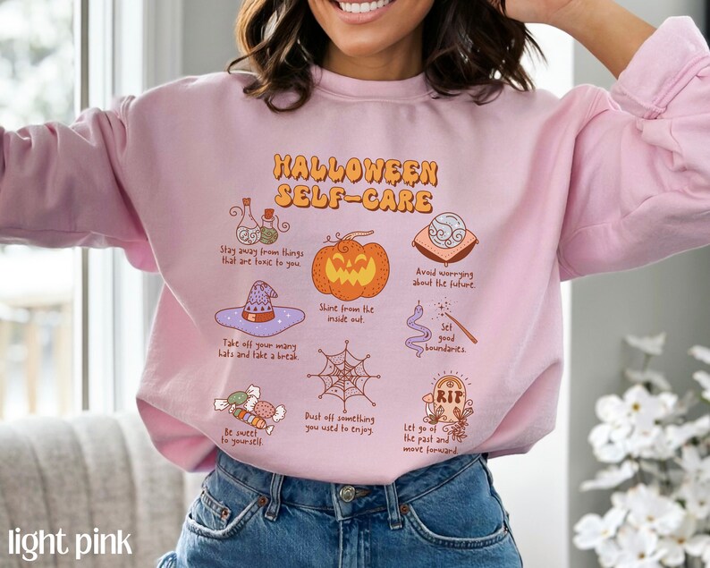 May include: A light pink sweatshirt with the text "HALLOWEEN SELF-CARE" in black lettering. The sweatshirt features a variety of Halloween-themed illustrations, including a pumpkin, a witch's hat, a spiderweb, and a tombstone.
