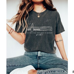 Puede incluir: Una camiseta gris oscuro con un gr&aacute;fico blanco de una tabla de surf y olas. El texto "Si las estrellas fueron hechas para adorar, yo tambi&eacute;n lo har&eacute;" est&aacute; impreso en la camiseta.