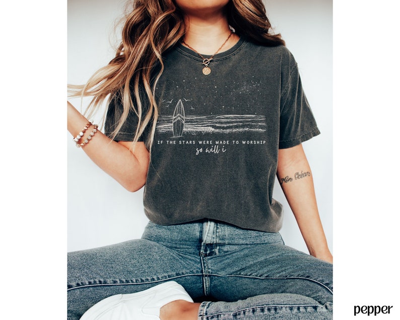 Puede incluir: Una camiseta gris oscuro con un gr&aacute;fico blanco de una tabla de surf y olas. El texto "Si las estrellas fueron hechas para adorar, yo tambi&eacute;n lo har&eacute;" est&aacute; impreso en la camiseta.