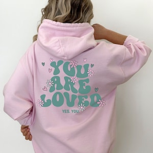 以下が含まれることがあります： 薄いピンク色のパーカー。 ターコイズ色の「YOU ARE LOVED」の文字が花とハートで囲まれています。「YES. YOU!」の文字が下にあります。 パーカーにはフードと長袖が付いています。