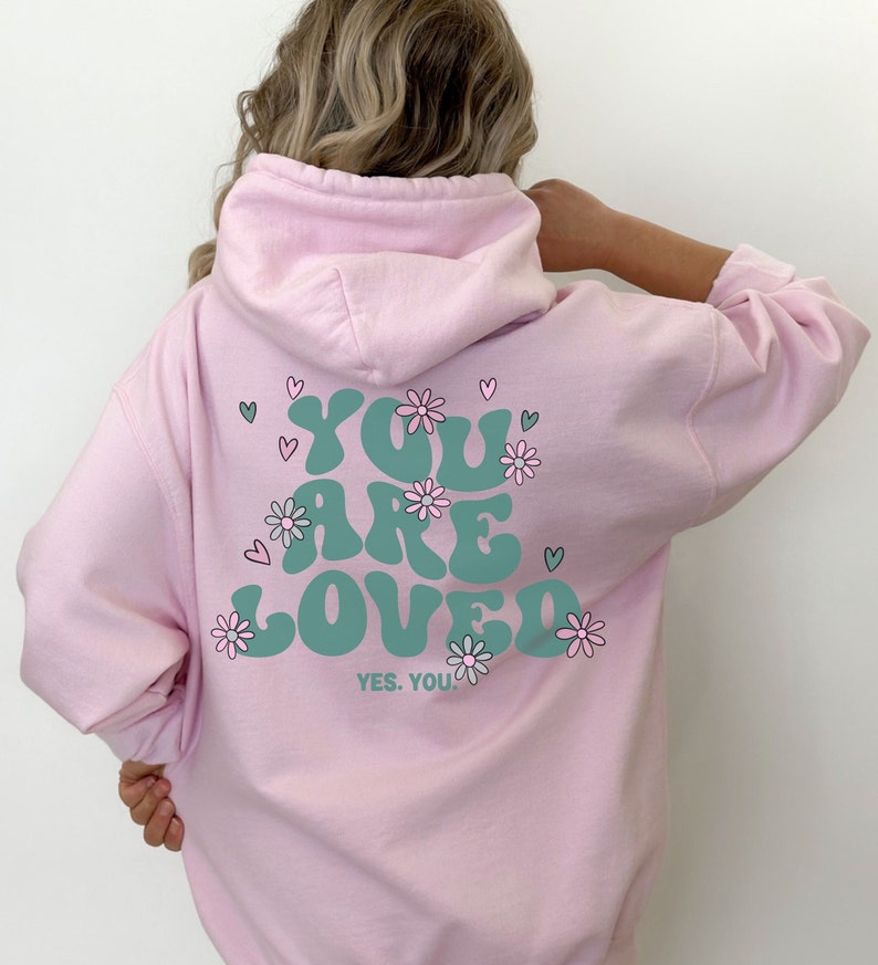以下が含まれることがあります： 薄いピンク色のパーカー。 ターコイズ色の「YOU ARE LOVED」の文字が花とハートで囲まれています。「YES. YOU!」の文字が下にあります。 パーカーにはフードと長袖が付いています。