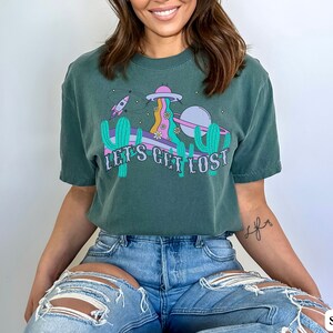 May include: A teal t-shirt with a graphic design. The design includes a UFO, rocket ship, cacti, and the text "LET'S GET LOST." The design has purple, pink, and yellow accents. The shirt is made of a soft material.
