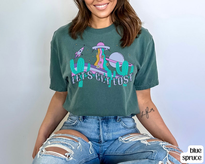 May include: A teal t-shirt with a graphic design. The design includes a UFO, rocket ship, cacti, and the text "LET'S GET LOST." The design has purple, pink, and yellow accents. The shirt is made of a soft material.