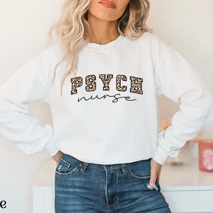 May include: White sweatshirt with the text "PSYCH nurse" in a combination of leopard print and script font. The word "white" is printed at the bottom left of the image. The sweatshirt is a crew neck style.