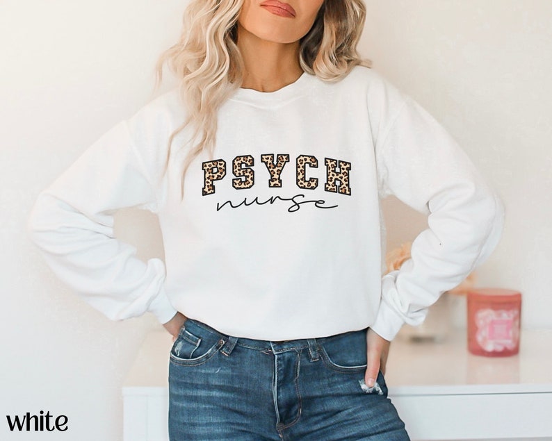 May include: White sweatshirt with the text "PSYCH nurse" in a combination of leopard print and script font. The word "white" is printed at the bottom left of the image. The sweatshirt is a crew neck style.
