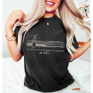 Puede incluir: Camiseta negra con un gr&aacute;fico blanco de una tabla de surf en una playa con estrellas en el cielo. El texto "If the stars were made to worship so will I" est&aacute; impreso en la camiseta.