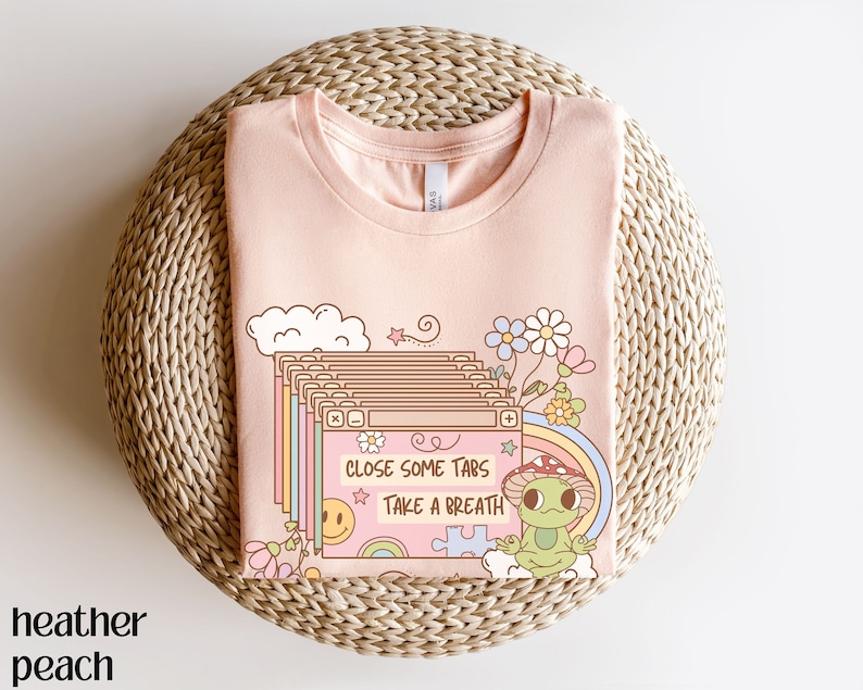 May include: A pink t-shirt with a cartoon illustration of a frog meditating in front of a computer screen. The text on the screen reads "Close some tabs take a breath".