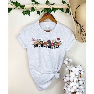 May include: A grey t-shirt with a floral and mushroom design and the word "NEUROSCIENCE" in black lettering.