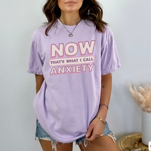 May include: A light purple t-shirt with the text "NOW THAT'S WHAT I CALL ANXIETY" in pink and white. The shirt is a relaxed fit and has short sleeves. The shirt is made of a soft material.