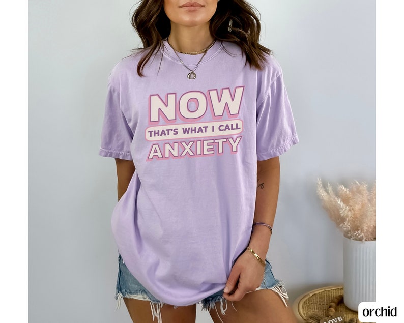 May include: A light purple t-shirt with the text "NOW THAT'S WHAT I CALL ANXIETY" in pink and white. The shirt is a relaxed fit and has short sleeves. The shirt is made of a soft material.