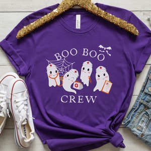 May include: Purple short-sleeved t-shirt with a graphic of three ghosts wearing nurse hats and holding medical supplies. The text "Boo Boo Crew" is printed below the ghosts.