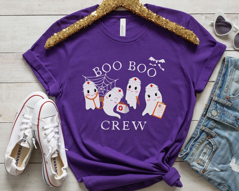May include: Purple short-sleeved t-shirt with a graphic of three ghosts wearing nurse hats and holding medical supplies. The text "Boo Boo Crew" is printed below the ghosts.