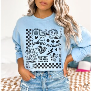 May include: Light blue sweatshirt with a graphic print of a sunflower, a rainbow, a sun, a lightning bolt, a butterfly, and a checkered pattern. The text on the sweatshirt reads "Be your own sunshine", "healing is not linear", "Find joy in the journey", "Don't let the hard days win", and "Love yourself".