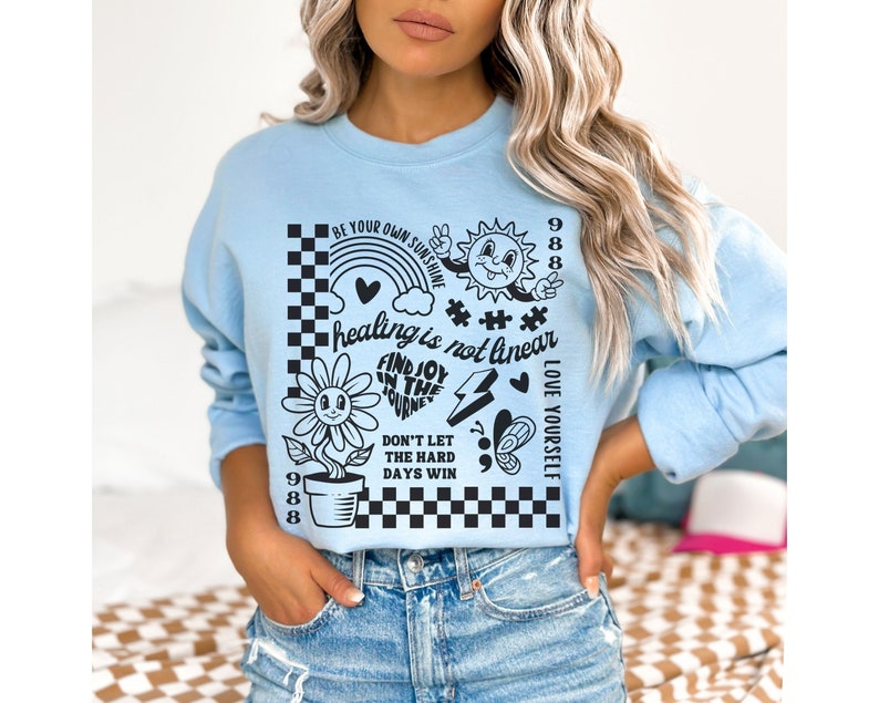 May include: Light blue sweatshirt with a graphic print of a sunflower, a rainbow, a sun, a lightning bolt, a butterfly, and a checkered pattern. The text on the sweatshirt reads "Be your own sunshine", "healing is not linear", "Find joy in the journey", "Don't let the hard days win", and "Love yourself".