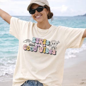May include: Cream-colored t-shirt with the text "SSRS and Good Vibes" in a retro font. The design includes a rainbow, a smiling sun, and other colorful elements. The shirt is a relaxed fit.
