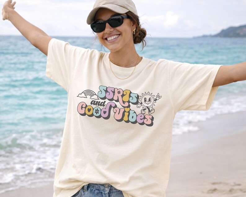 May include: Cream-colored t-shirt with the text "SSRS and Good Vibes" in a retro font. The design includes a rainbow, a smiling sun, and other colorful elements. The shirt is a relaxed fit.