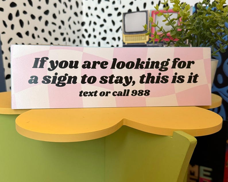 Puede incluir: Un letrero rectangular con el texto "If you are looking for a sign to stay, this is it" en fuente negra. El fondo tiene un patr&oacute;n de cuadros rosas y blancos. El texto "text or call 988" est&aacute; debajo.