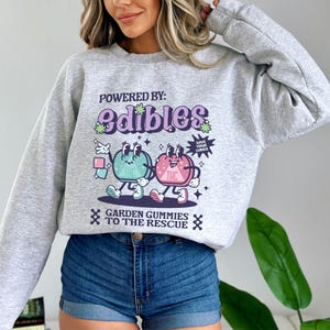 May include: Gray sweatshirt with a graphic print of two cartoon gummies. The gummies are green and pink and have faces. The text on the sweatshirt says "Powered by: Edibles" and "Garden Gummies to the Rescue".