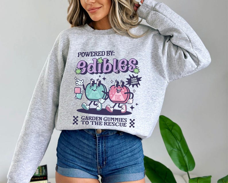 May include: Gray sweatshirt with a graphic print of two cartoon gummies. The gummies are green and pink and have faces. The text on the sweatshirt says "Powered by: Edibles" and "Garden Gummies to the Rescue".