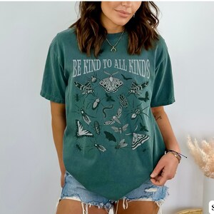 Op de afbeelding: Een blauwgroen t-shirt met de tekst "BE KIND TO ALL KINDS" in een vintage lettertype. Het shirt toont illustraties van verschillende insecten en vlinders in grijstinten.