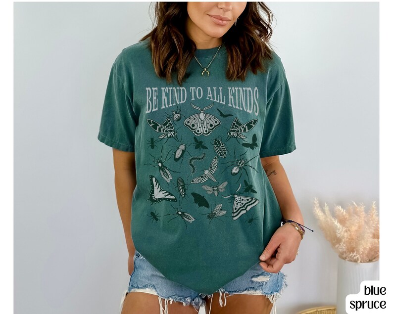 Op de afbeelding: Een blauwgroen t-shirt met de tekst "BE KIND TO ALL KINDS" in een vintage lettertype. Het shirt toont illustraties van verschillende insecten en vlinders in grijstinten.