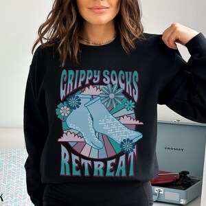 May include: Black sweatshirt with a graphic design of blue socks with a retro-style design and the text "Grippy Socks Retreat".