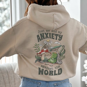 May include: A beige hoodie with a graphic print of a frog sitting on a mushroom. The graphic also includes a snail, a daisy, and the text "Just me and my anxiety against the world".