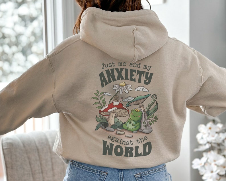 May include: A beige hoodie with a graphic print of a frog sitting on a mushroom. The graphic also includes a snail, a daisy, and the text "Just me and my anxiety against the world".
