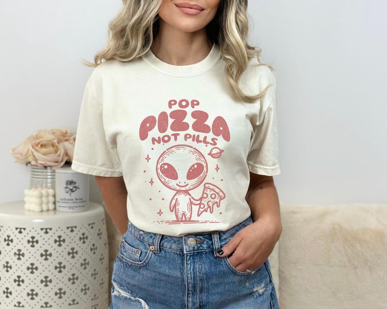 May include: A white t-shirt with a pink alien graphic holding a slice of pizza. The text on the shirt reads "Pop Pizza Not Pills".