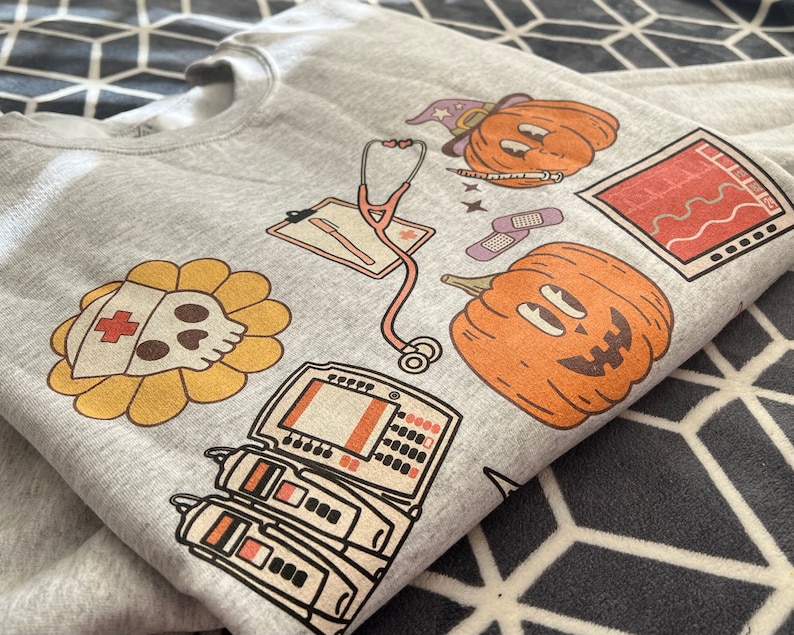 May include: A gray sweatshirt with a Halloween-themed medical design. It features a pumpkin with a witch's hat, a skull with a nurse's cap, a stethoscope, a medical monitor, and a heart rate monitor.