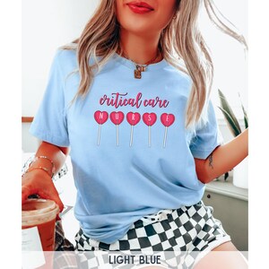 Pu&ograve; includere: Maglietta blu chiaro con il testo "critical care nurse" e lecca-lecca a forma di cuore rosa con le lettere "nurse" su di esse.