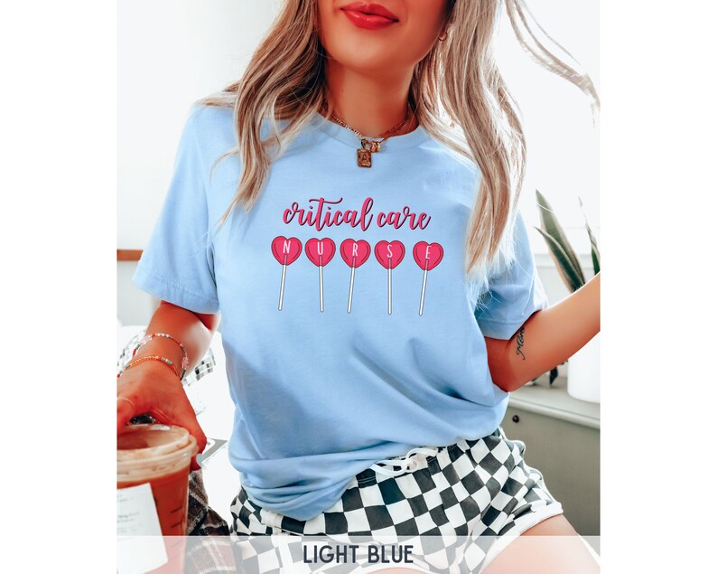 Pu&ograve; includere: Maglietta blu chiaro con il testo "critical care nurse" e lecca-lecca a forma di cuore rosa con le lettere "nurse" su di esse.