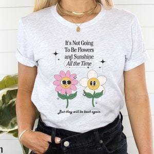 May include: A white t-shirt with a graphic of two cartoon flowers with faces. The text on the shirt reads "It's Not Going To Be Flowers and Sunshine All the Time But they will be back again".