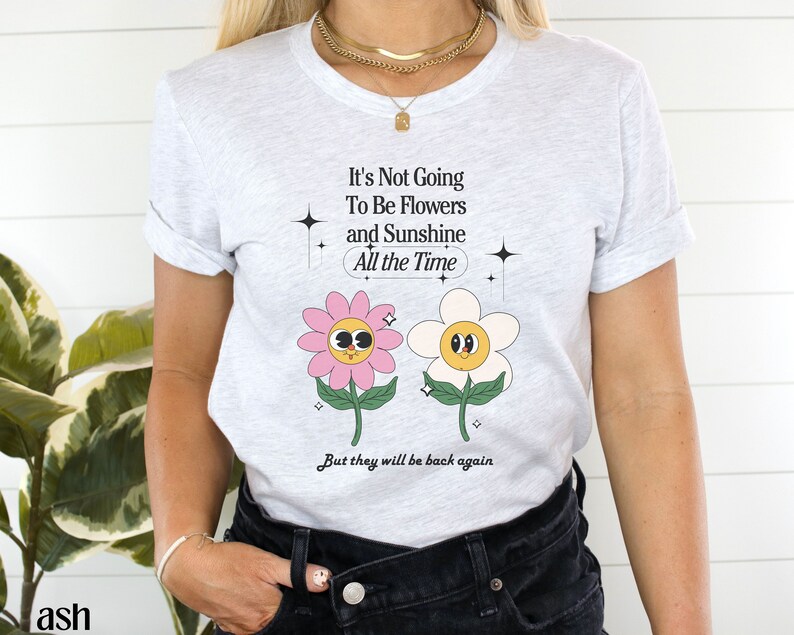 May include: A white t-shirt with a graphic of two cartoon flowers with faces. The text on the shirt reads "It's Not Going To Be Flowers and Sunshine All the Time But they will be back again".