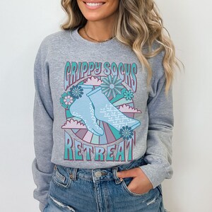 May include: Gray sweatshirt with a retro-style graphic of blue grippy socks with a floral design and the text "Grippy Socks Retreat".