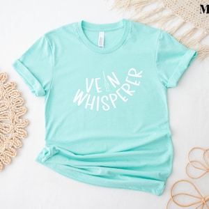 以下が含まれることがあります： 薄い青色のTシャツに、白い文字で「Vein Whisperer」と書いてあります。テキストは、中央に小さな針と注射器のグラフィックがある円弧の形をしています。