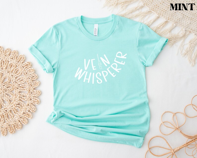 以下が含まれることがあります： 薄い青色のTシャツに、白い文字で「Vein Whisperer」と書いてあります。テキストは、中央に小さな針と注射器のグラフィックがある円弧の形をしています。