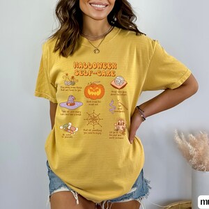 May include: A yellow t-shirt with a graphic print of a Halloween self-care guide. The graphic includes illustrations of a witch's hat, a pumpkin, a spiderweb, and other Halloween-themed items. The text on the shirt reads "Halloween Self-Care".