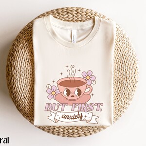 May include: A beige t-shirt with a graphic of a pink coffee cup with a face and the text "But First, Anxiety".