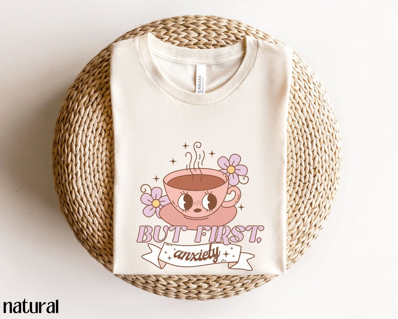 May include: A beige t-shirt with a graphic of a pink coffee cup with a face and the text "But First, Anxiety".