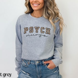 May include: A light grey sweatshirt with the text "PSYCH nurse" in a combination of leopard print and script font. The sweatshirt is a crew neck style and has long sleeves. The words "sport grey" are at the bottom of the image.